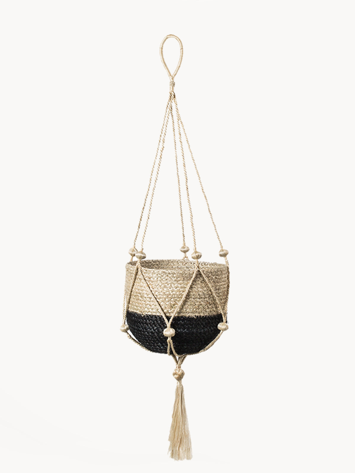 The Plant Hanger - Nadu features a woven basket with a black and natural fiber design, suspended by a handmade jute hanger adorned with braided and knotted ropes, complete with a tassel at the bottom.