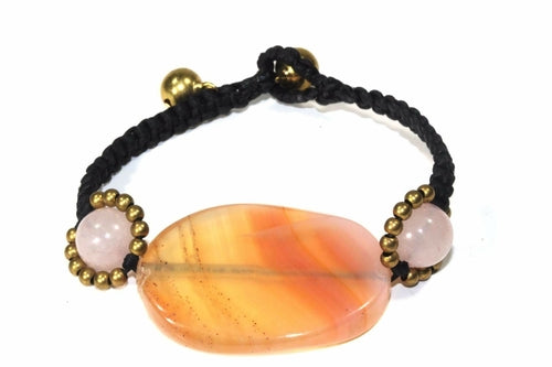 Two hands, adorned with handmade jewelry featuring a large oval stone and floral designs, gracefully hold a pink hibiscus against patterned fabric. One bracelet, the Peach Agate Slice & Rose Quartz, showcases delicate rose quartz rounds, enhancing the scene's elegance.