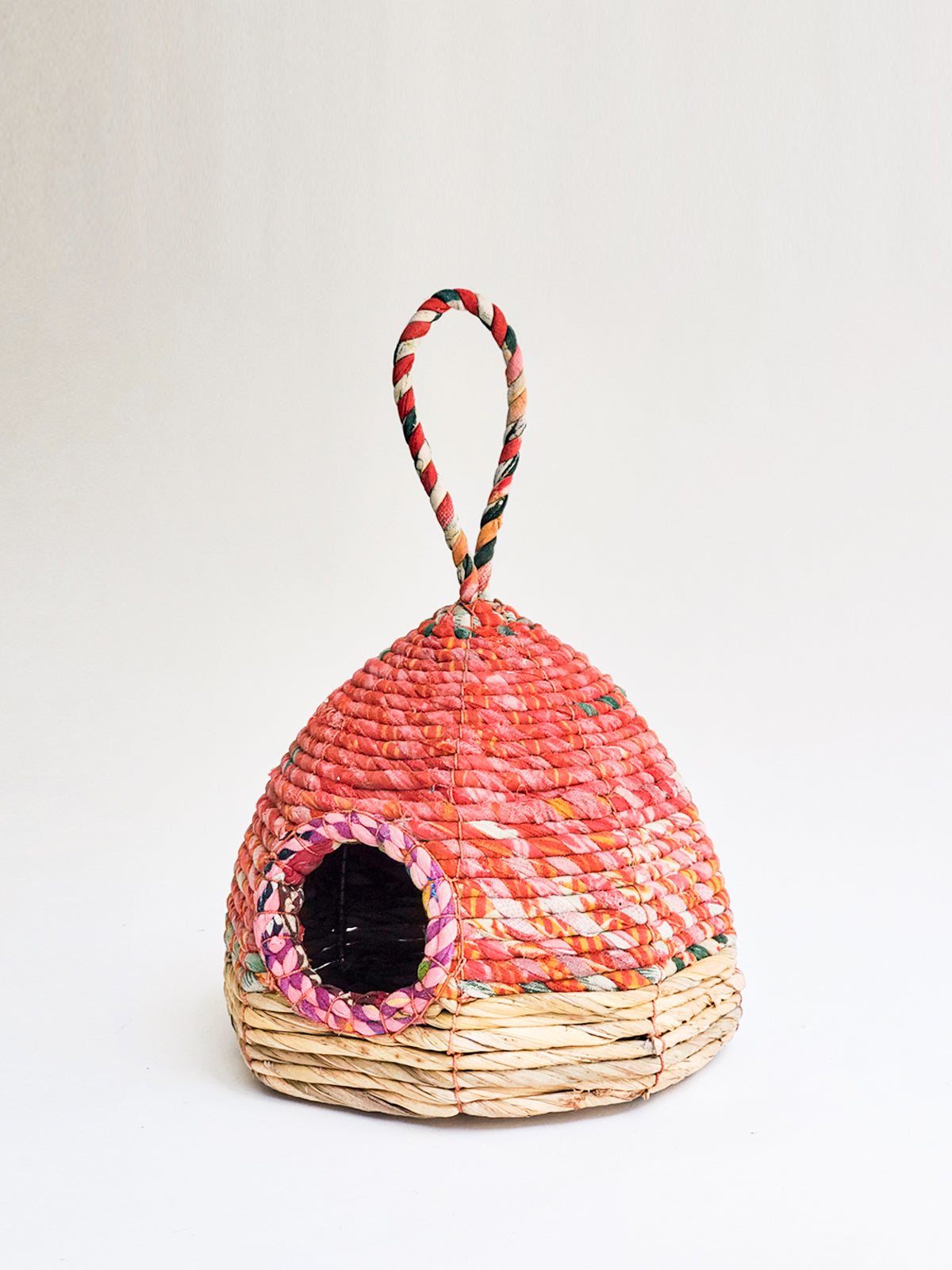 The Seagrass & Sari Birdhouse - Garden is an eco-friendly, handcrafted birdhouse showcasing a woven red and beige design, featuring a circular entrance and a looped handle on top.