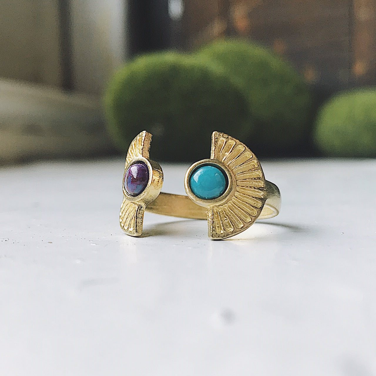 Two artistic rings are displayed on a white surface, one exuding bohemian charm with its gold band and captivating copper turquoise stone, while the other showcases a silver band paired with a dark purple stone. Both pieces from the "Dawn to Dusk - Two Stone Turquoise Boho Sunburst Ring" collection radiate galaxy flair and offer the convenience of an adjustable design.