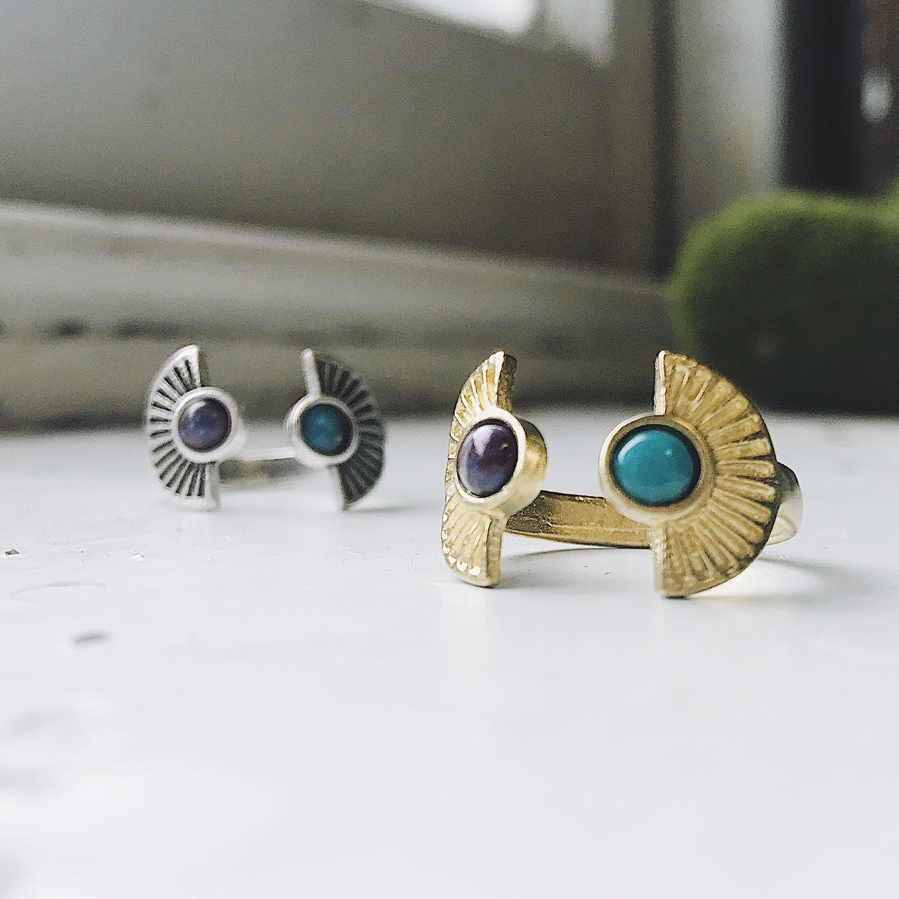 Two artistic rings are displayed on a white surface, one exuding bohemian charm with its gold band and captivating copper turquoise stone, while the other showcases a silver band paired with a dark purple stone. Both pieces from the "Dawn to Dusk - Two Stone Turquoise Boho Sunburst Ring" collection radiate galaxy flair and offer the convenience of an adjustable design.