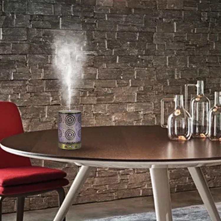 A chic Color Changing Aromatherapy Oil Diffuser with a floral motif rests on a wooden table, softly releasing mist enhanced with essential oils. In the background, a vase displays vibrant flowers, crafting an ideal aromatherapy atmosphere.