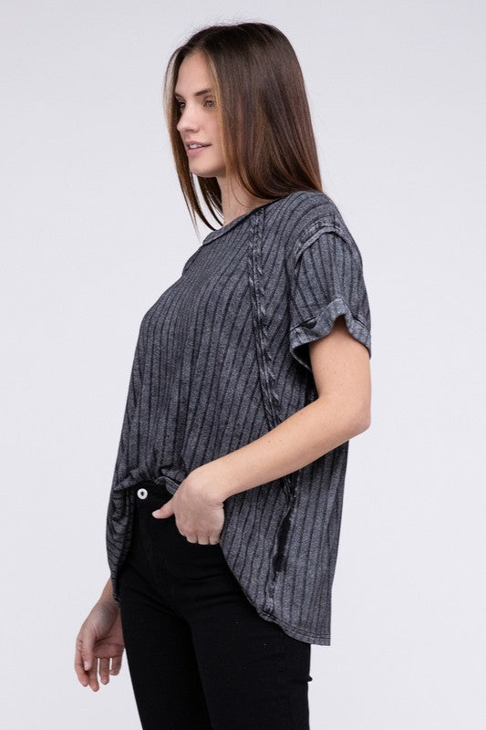 A woman in a Ribbed Raglan Dolman Sleeve Boat-Neck Top and white pants, standing against a gray background, gently pulling the fabric of her top.