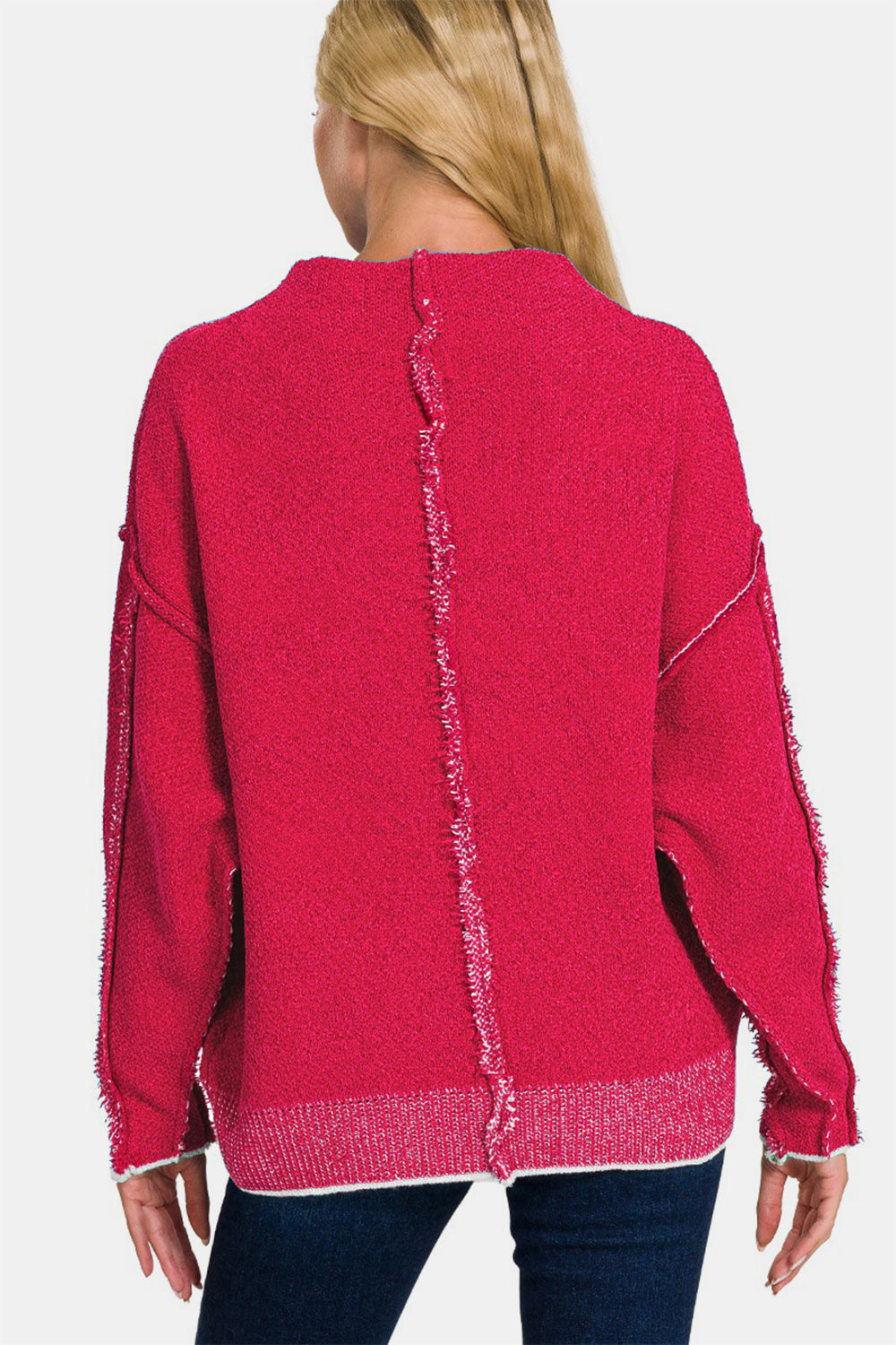 A woman in a vibrant pink Zenana Exposed Seam Mock Neck Long Sleeve Sweater and jeans stands elegantly against a minimalist white background.