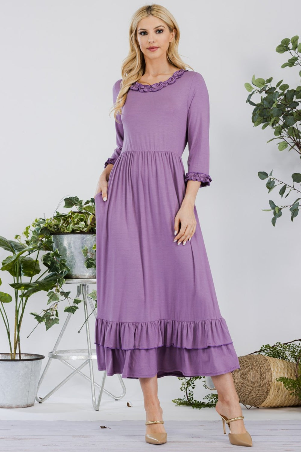 A woman in the Celeste Full Size Layered Ruffle Hem Dress with Pockets, featuring an A-line silhouette and 3/4 sleeves, stands on a wooden floor surrounded by potted plants and a wicker basket.