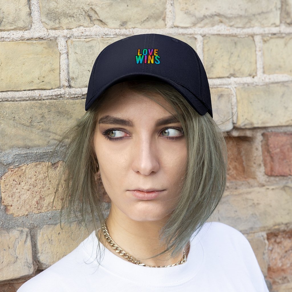 A bearded man wears the Rainbow Love Wins Unisex Twill Cap adorned with the phrase "Rainbow Love Wins" in vibrant colors, along with a gray sweatshirt, as he gazes into the distance against a simple backdrop.
