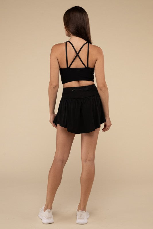 Close-up of a woman in a black crop top and Wide Band Tennis Skirt with Zippered Back Pocket, pulling the skirt to show matching shorts with a zippered back pocket underneath.
