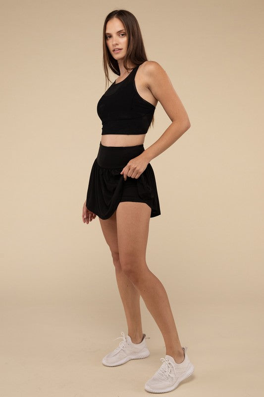 Close-up of a woman in a black crop top and Wide Band Tennis Skirt with Zippered Back Pocket, pulling the skirt to show matching shorts with a zippered back pocket underneath.