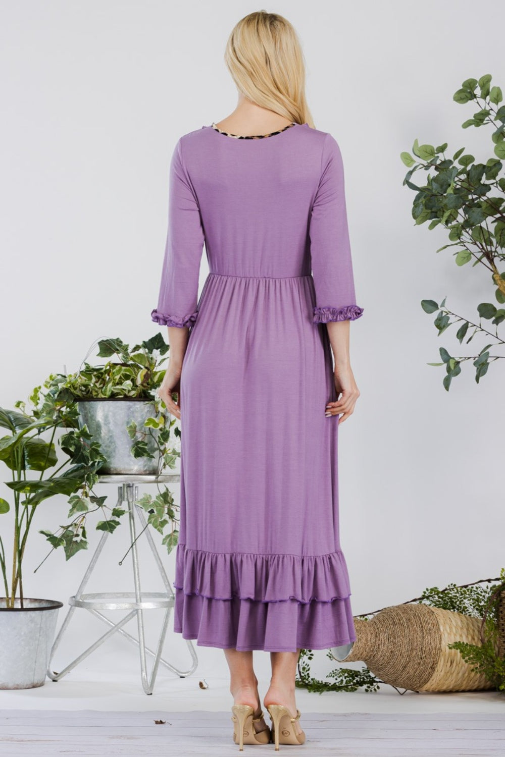 A woman in the Celeste Full Size Layered Ruffle Hem Dress with Pockets, featuring an A-line silhouette and 3/4 sleeves, stands on a wooden floor surrounded by potted plants and a wicker basket.