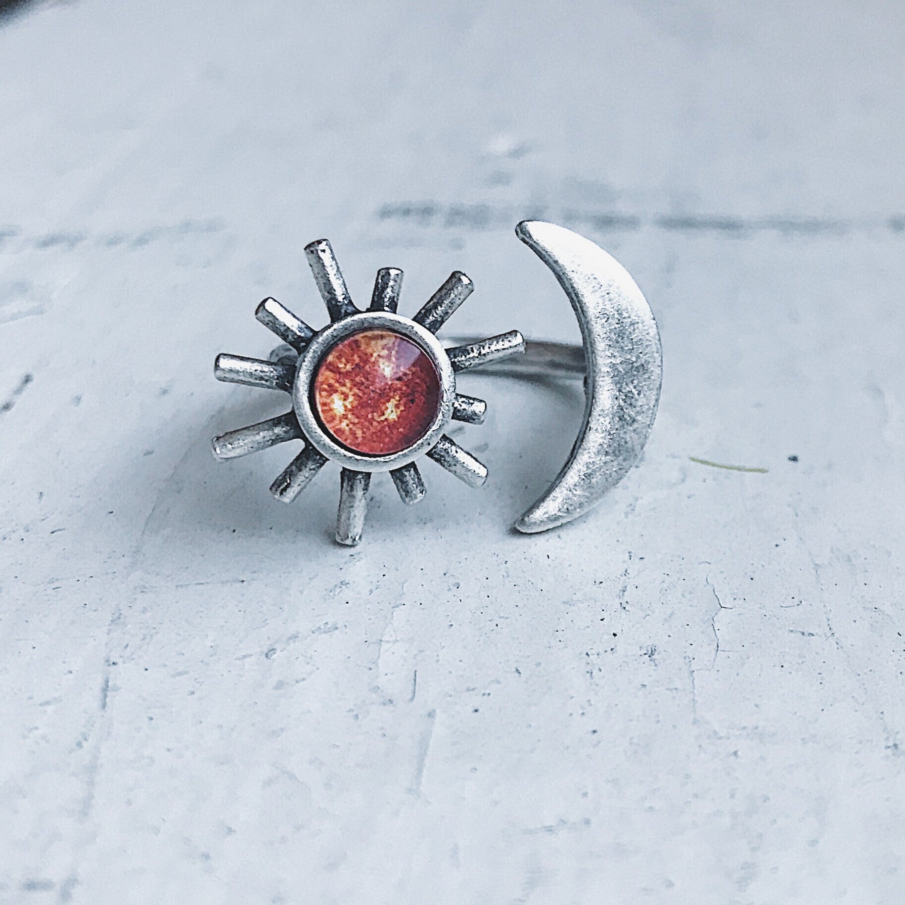 A hand adorned with the Sun and Moon Sculptural Statement Ring, showcasing a celestial design of a crescent moon and sun paired with a captivating red-orange gemstone. This adjustable ring beautifully captures the enchanting dance of day and night.