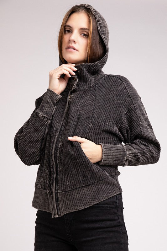 Woman in a Acid Wash Cotton Waffle Hooded Zip-Up Jacket with pockets and black pants, smiling and posing with hands on hips, against a light gray background.