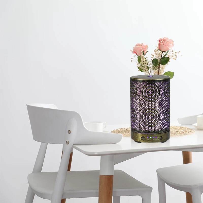 A chic Color Changing Aromatherapy Oil Diffuser with a floral motif rests on a wooden table, softly releasing mist enhanced with essential oils. In the background, a vase displays vibrant flowers, crafting an ideal aromatherapy atmosphere.