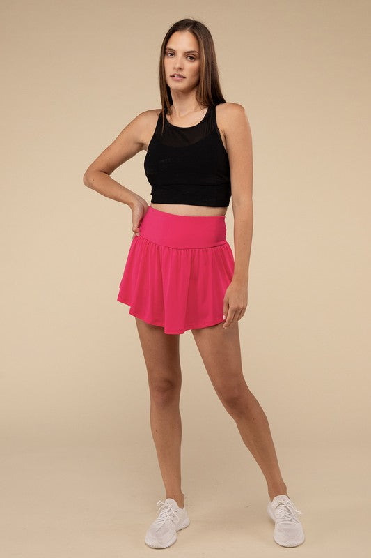 Close-up of a woman in a black crop top and Wide Band Tennis Skirt with Zippered Back Pocket, pulling the skirt to show matching shorts with a zippered back pocket underneath.