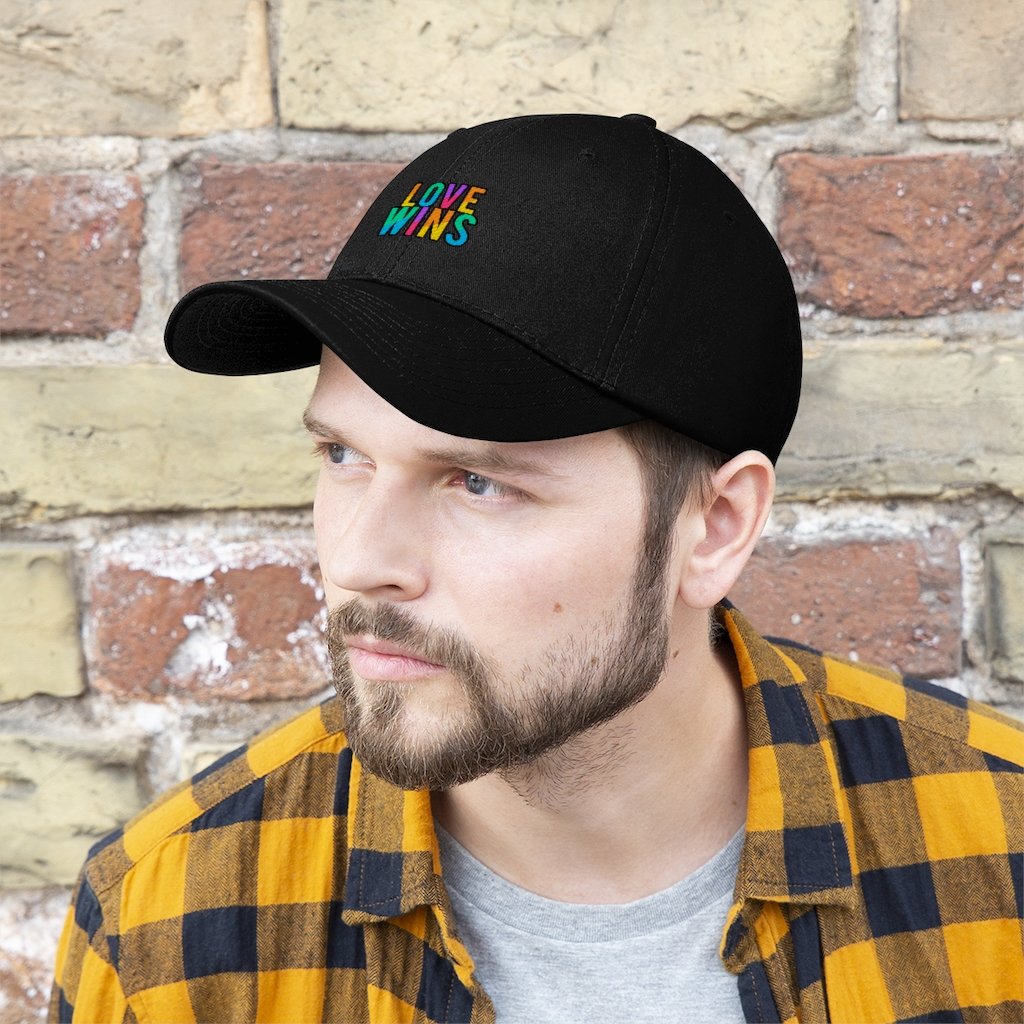 A bearded man wears the Rainbow Love Wins Unisex Twill Cap adorned with the phrase "Rainbow Love Wins" in vibrant colors, along with a gray sweatshirt, as he gazes into the distance against a simple backdrop.