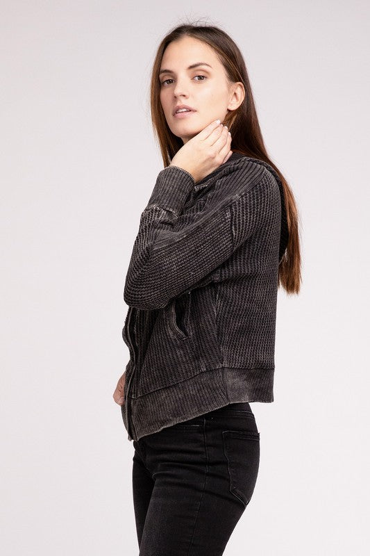 Woman in a Acid Wash Cotton Waffle Hooded Zip-Up Jacket with pockets and black pants, smiling and posing with hands on hips, against a light gray background.