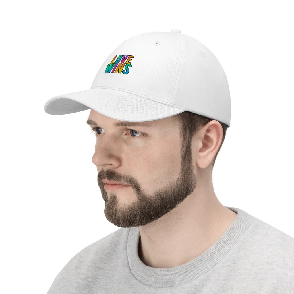 A bearded man wears the Rainbow Love Wins Unisex Twill Cap adorned with the phrase "Rainbow Love Wins" in vibrant colors, along with a gray sweatshirt, as he gazes into the distance against a simple backdrop.
