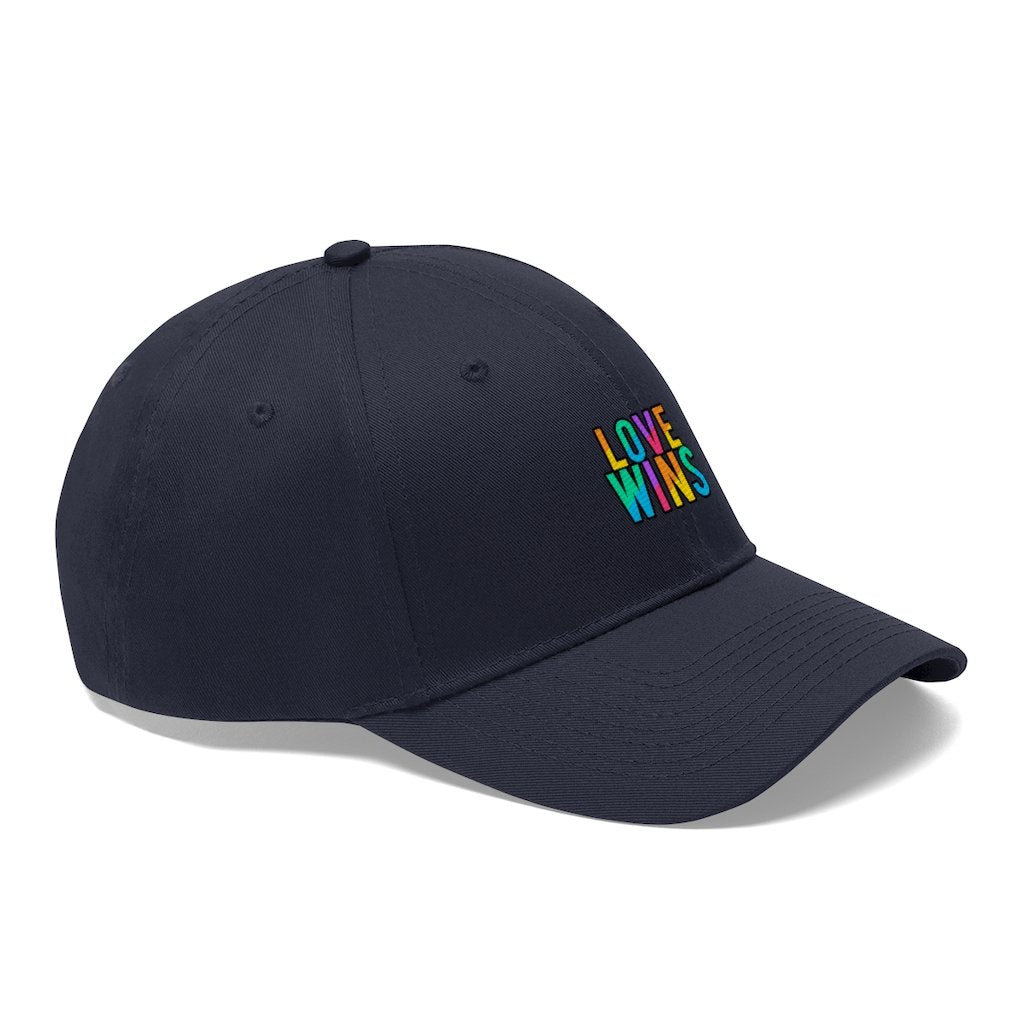 A bearded man wears the Rainbow Love Wins Unisex Twill Cap adorned with the phrase "Rainbow Love Wins" in vibrant colors, along with a gray sweatshirt, as he gazes into the distance against a simple backdrop.