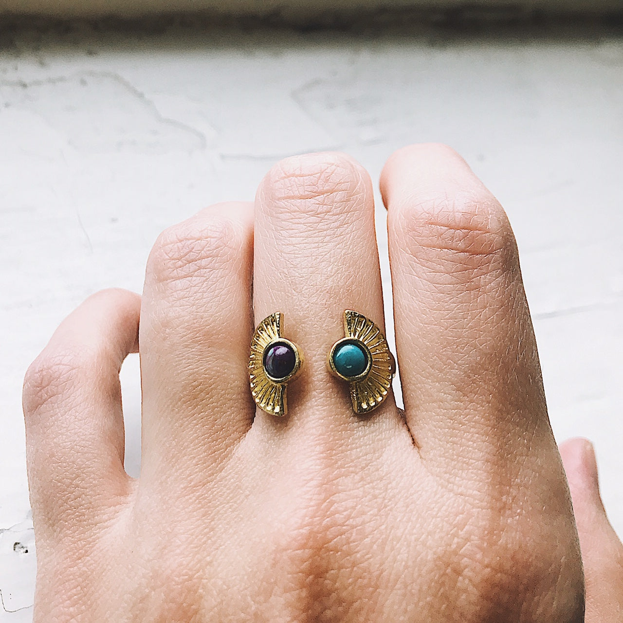 Two artistic rings are displayed on a white surface, one exuding bohemian charm with its gold band and captivating copper turquoise stone, while the other showcases a silver band paired with a dark purple stone. Both pieces from the "Dawn to Dusk - Two Stone Turquoise Boho Sunburst Ring" collection radiate galaxy flair and offer the convenience of an adjustable design.