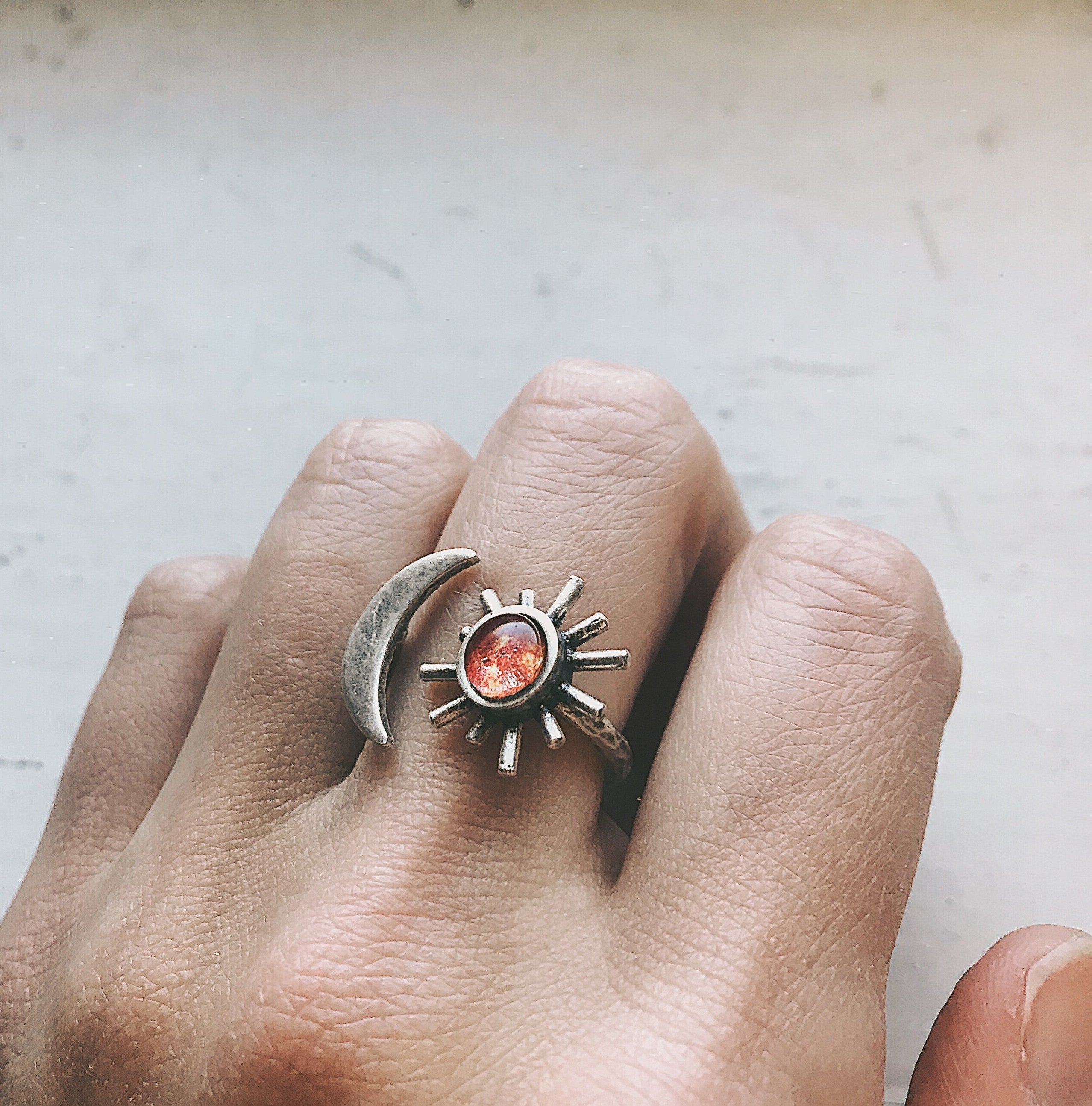 A hand adorned with the Sun and Moon Sculptural Statement Ring, showcasing a celestial design of a crescent moon and sun paired with a captivating red-orange gemstone. This adjustable ring beautifully captures the enchanting dance of day and night.