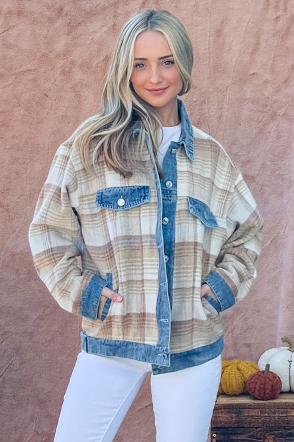 Dressed in the And The Why Full Size Washed Denim Detail Brushed Plaid Jacket and white pants, a person stands against a textured background, exuding vintage charm while surrounded by small decorative pumpkins.