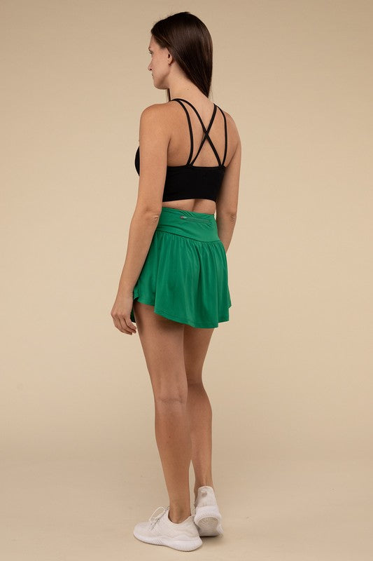 Close-up of a woman in a black crop top and Wide Band Tennis Skirt with Zippered Back Pocket, pulling the skirt to show matching shorts with a zippered back pocket underneath.