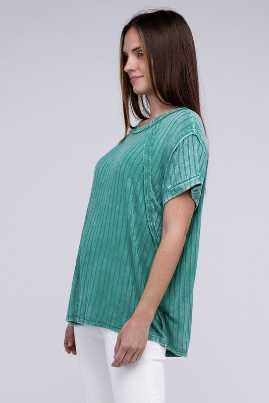 A woman in a Ribbed Raglan Dolman Sleeve Boat-Neck Top and white pants, standing against a gray background, gently pulling the fabric of her top.