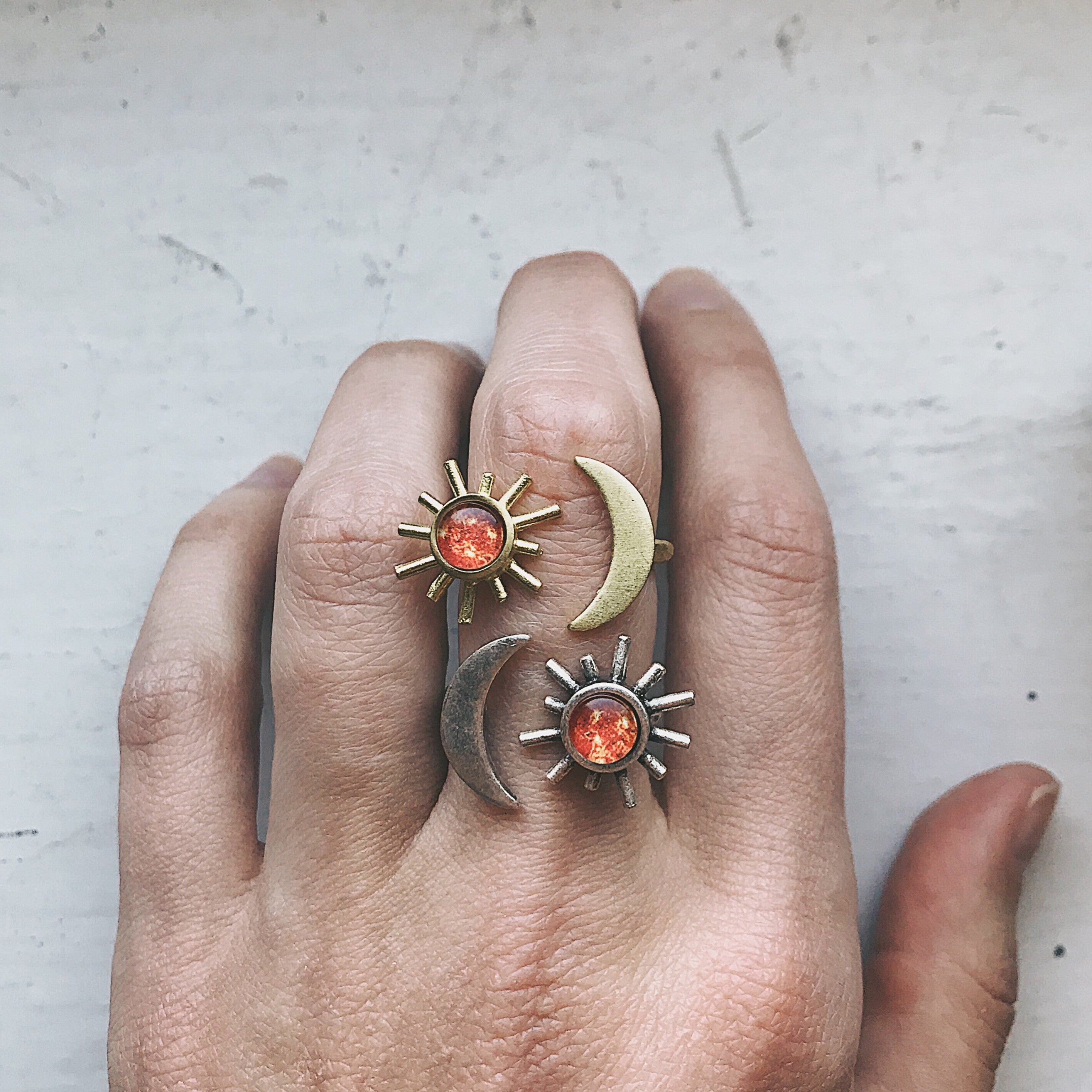 A hand adorned with the Sun and Moon Sculptural Statement Ring, showcasing a celestial design of a crescent moon and sun paired with a captivating red-orange gemstone. This adjustable ring beautifully captures the enchanting dance of day and night.
