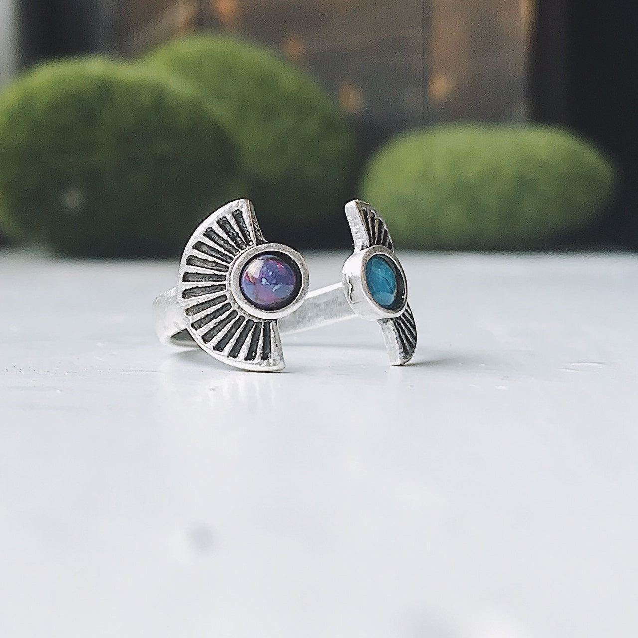 Two artistic rings are displayed on a white surface, one exuding bohemian charm with its gold band and captivating copper turquoise stone, while the other showcases a silver band paired with a dark purple stone. Both pieces from the "Dawn to Dusk - Two Stone Turquoise Boho Sunburst Ring" collection radiate galaxy flair and offer the convenience of an adjustable design.
