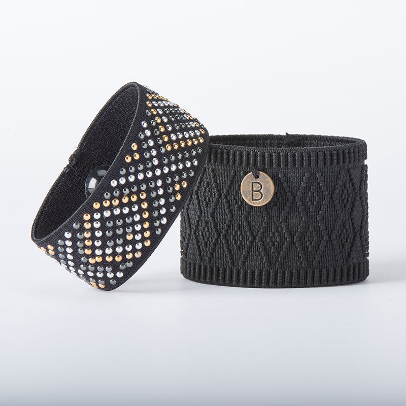 Two Cecilia + Aria Nausea Relief Bracelets feature black fabric wristbands: one adorned with metallic studs in a diamond pattern, and the other with a textured design and a small "B" metal tag. Stylish and functional, these bands also serve as acupressure bracelets to alleviate nausea and motion sickness.