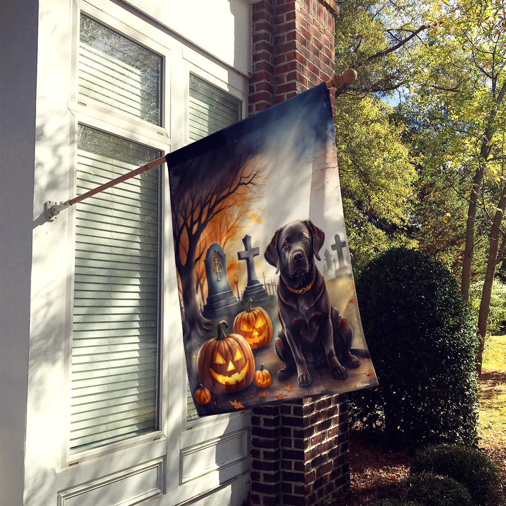 A Chocolate Labrador Retriever sits in a cemetery surrounded by carved pumpkins with glowing faces. Tombstones and bare trees loom in the background, while a Spooky Halloween House Flag featuring the dog waves gently, enhancing the spooky atmosphere.