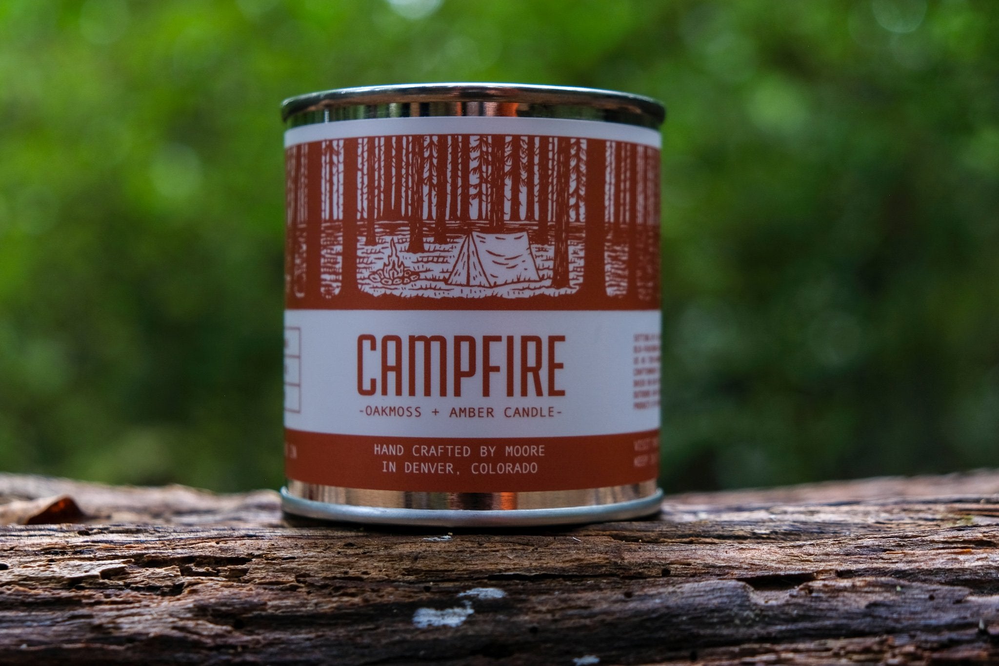 A tin candle called "Campfire Candle" sits on a log, featuring a forest illustration on its label. Handcrafted with natural soy by Moore in Denver, Colorado, this candle releases an inviting outdoor aroma. Lush greenery in the background adds to its rustic charm.