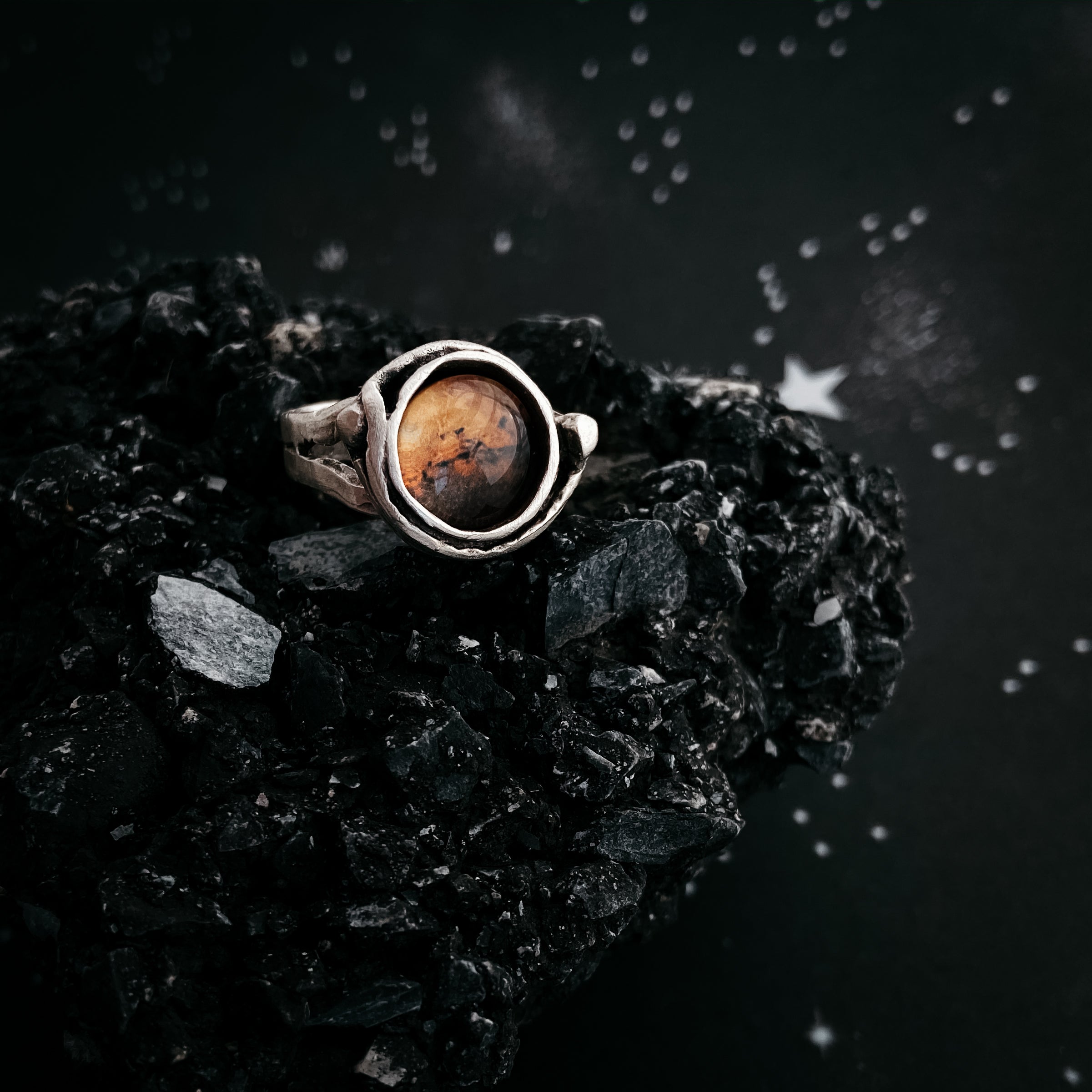 A hand wears the Mars and Moons Ring, featuring a silver tone with a spherical, galaxy-like design reminiscent of space exploration jewelry, set against a starry background.