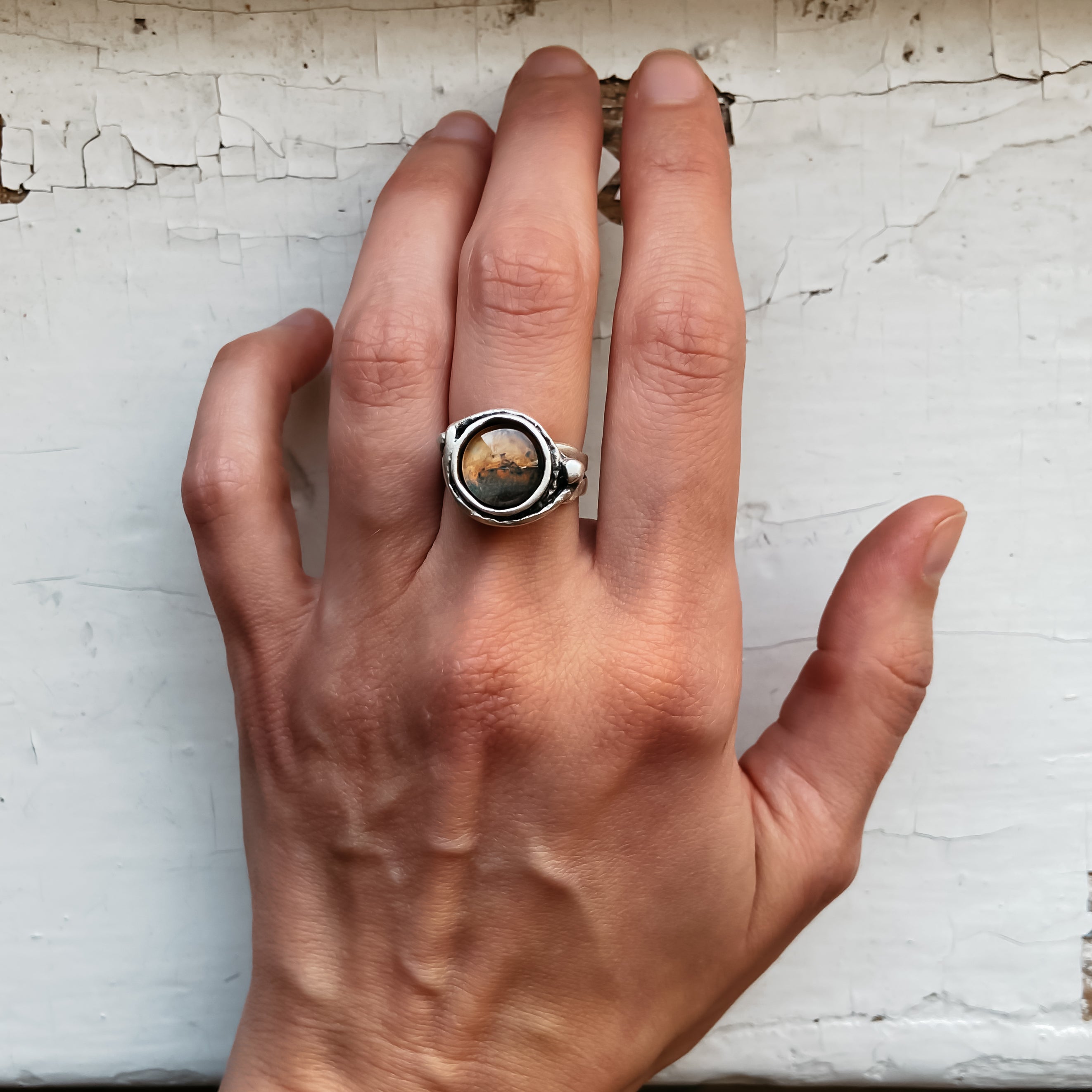 A hand wears the Mars and Moons Ring, featuring a silver tone with a spherical, galaxy-like design reminiscent of space exploration jewelry, set against a starry background.