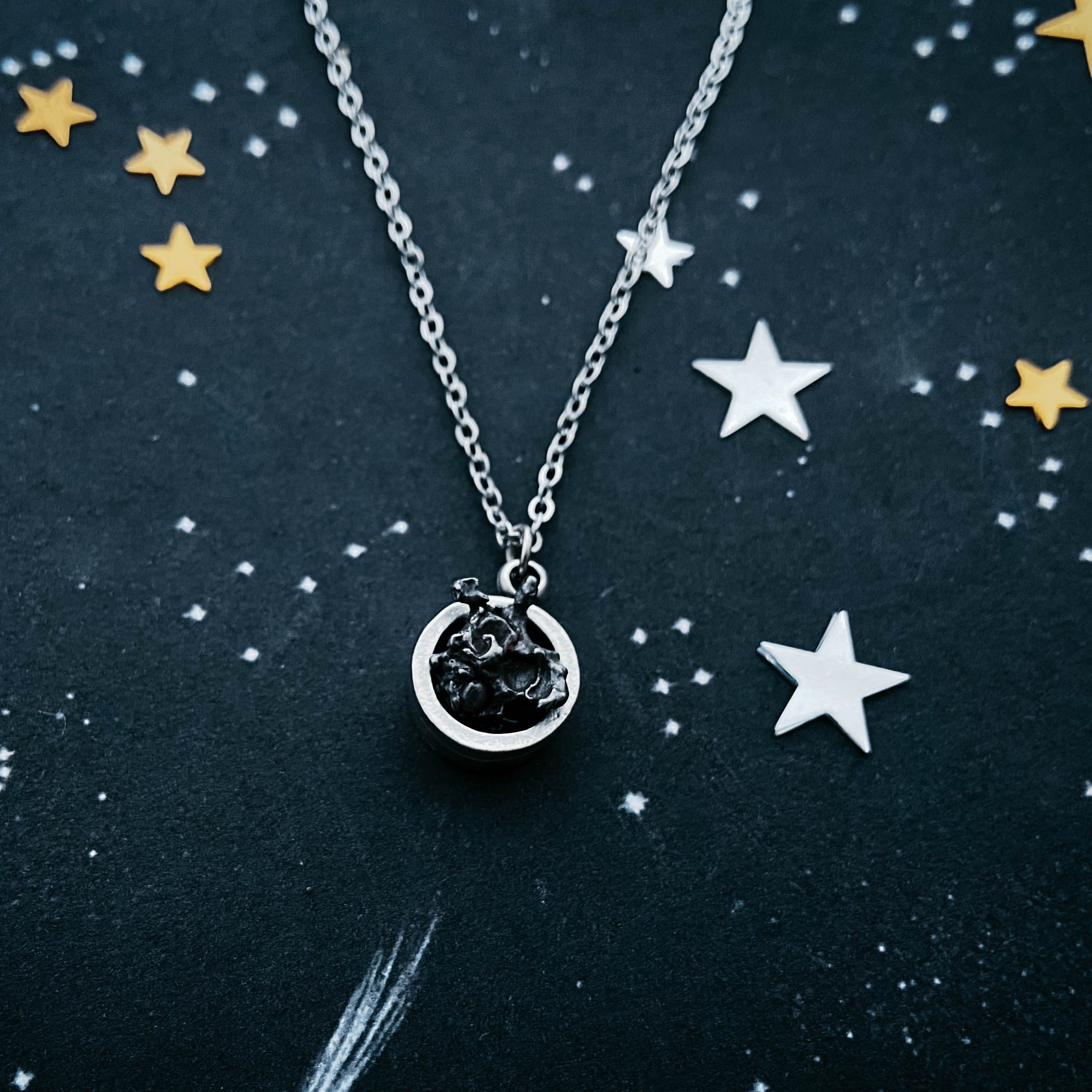 A Small Round Meteorite Pendant Necklace in Matte Brushed Silver rests on a dark background, surrounded by scattered gold and silver star-shaped decorations, embodying the enchanting allure of outer space jewelry.