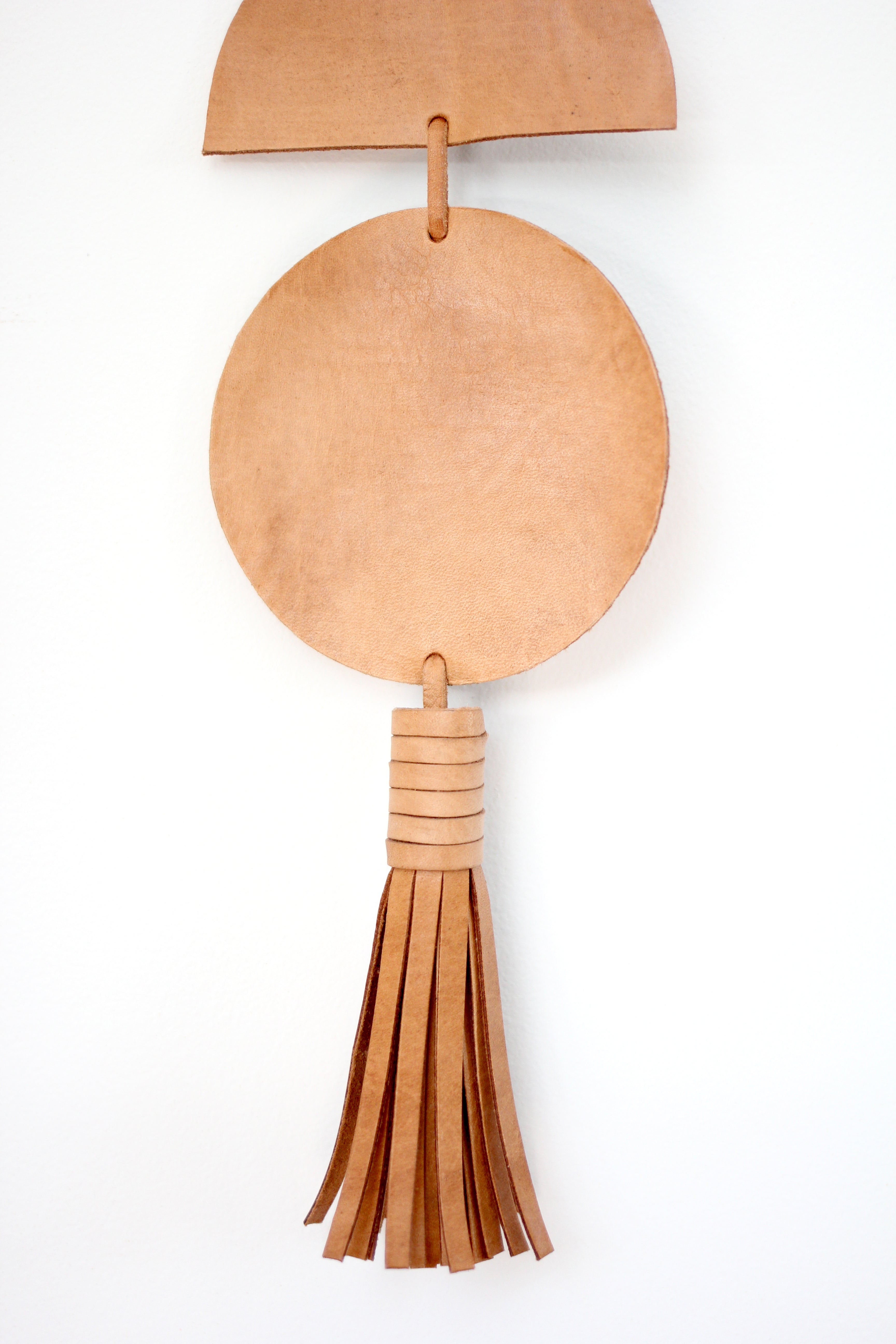 The Luna Moon Phase Wall Hanging, crafted with handmade circular and semicircular shapes from brown leather, is placed beside a large green monstera leaf on a white background, evoking the phases of the moon.