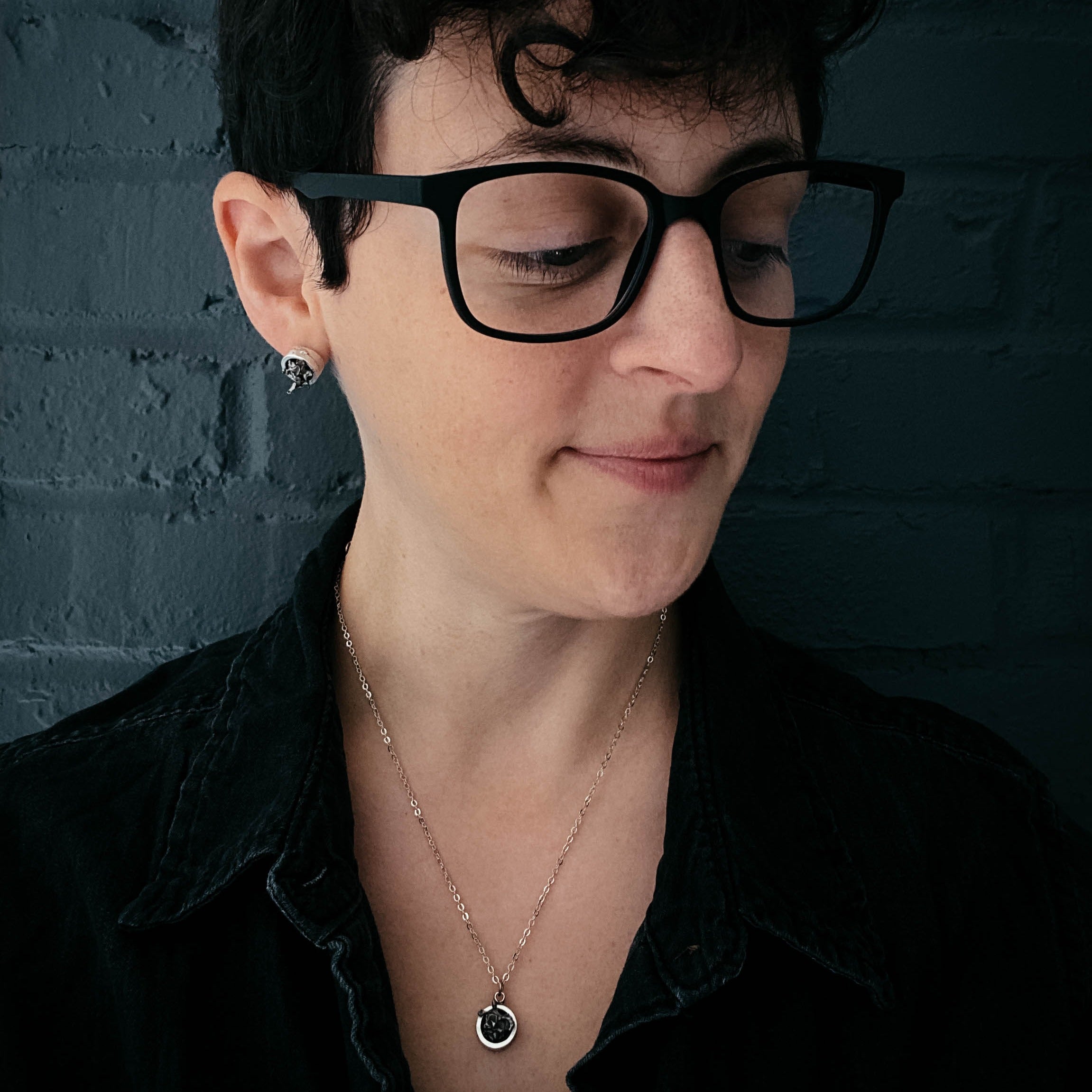 A Small Round Meteorite Pendant Necklace in Matte Brushed Silver rests on a dark background, surrounded by scattered gold and silver star-shaped decorations, embodying the enchanting allure of outer space jewelry.