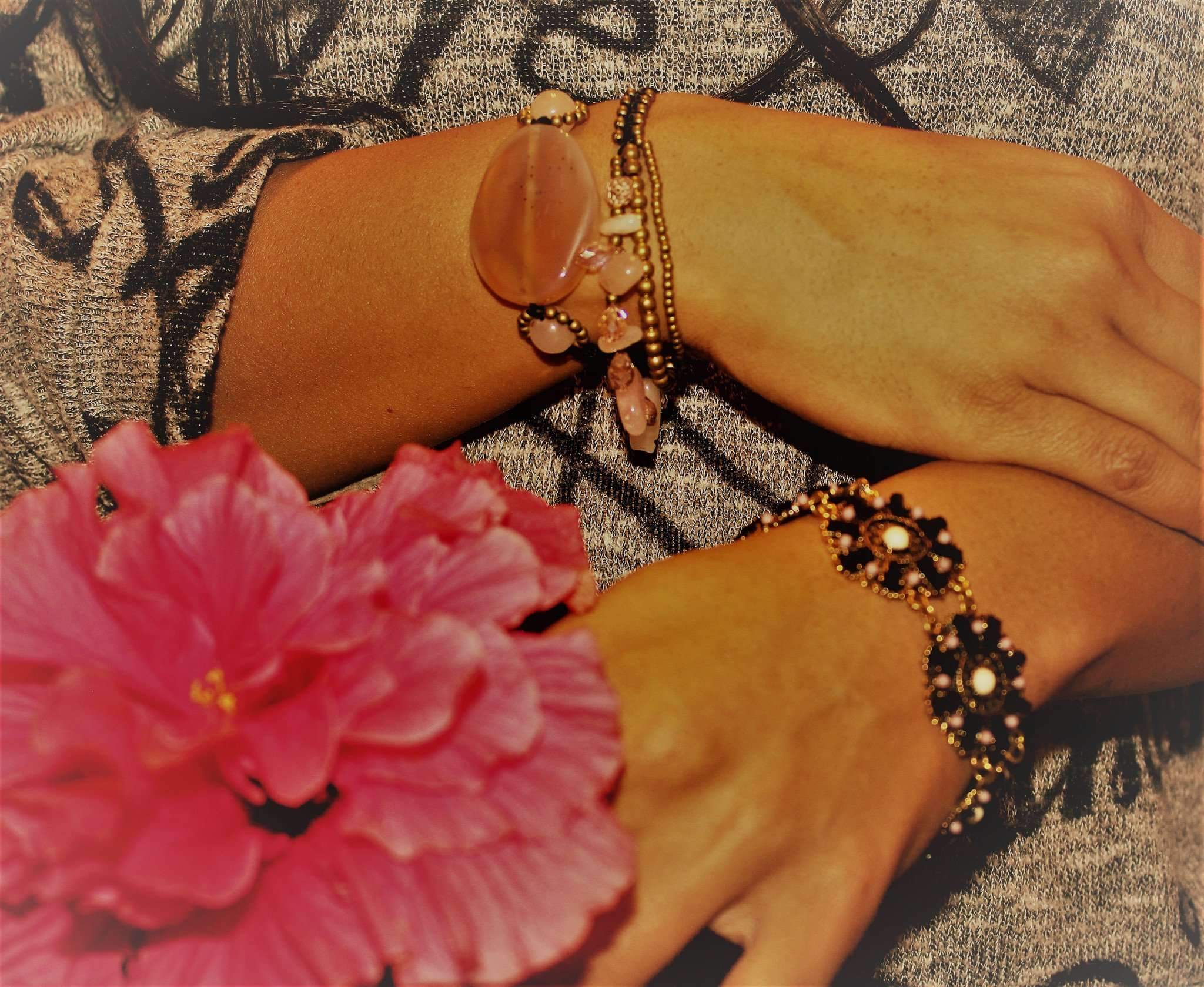 Two hands, adorned with handmade jewelry featuring a large oval stone and floral designs, gracefully hold a pink hibiscus against patterned fabric. One bracelet, the Peach Agate Slice & Rose Quartz, showcases delicate rose quartz rounds, enhancing the scene's elegance.
