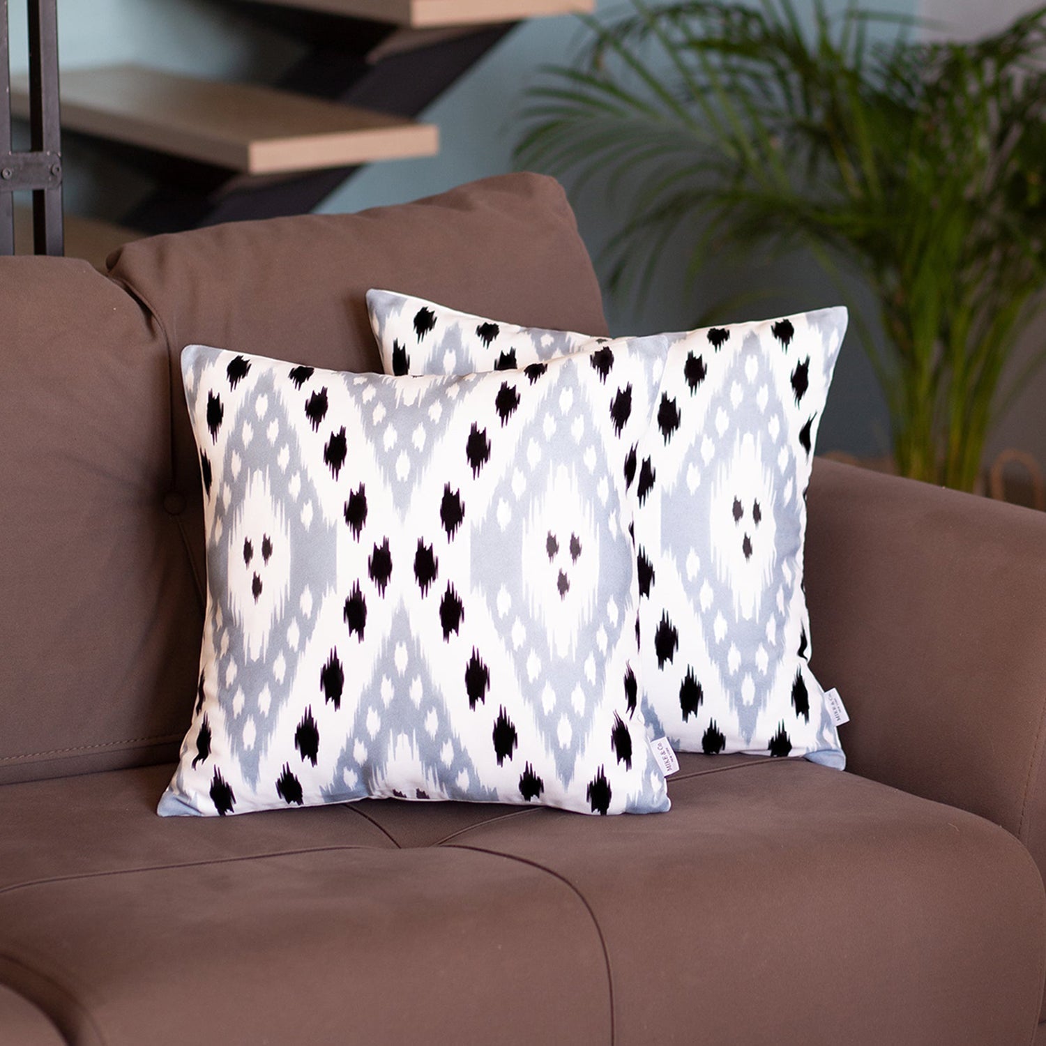 A pair of pillow covers from the Ikat Set of 2 Square 18’’x18’’ Boho Throw Pillow Covers, showcasing a blue and white ikat design with hidden zipper closures, are placed on a brown sofa. In the background, you can see a potted plant and a staircase.