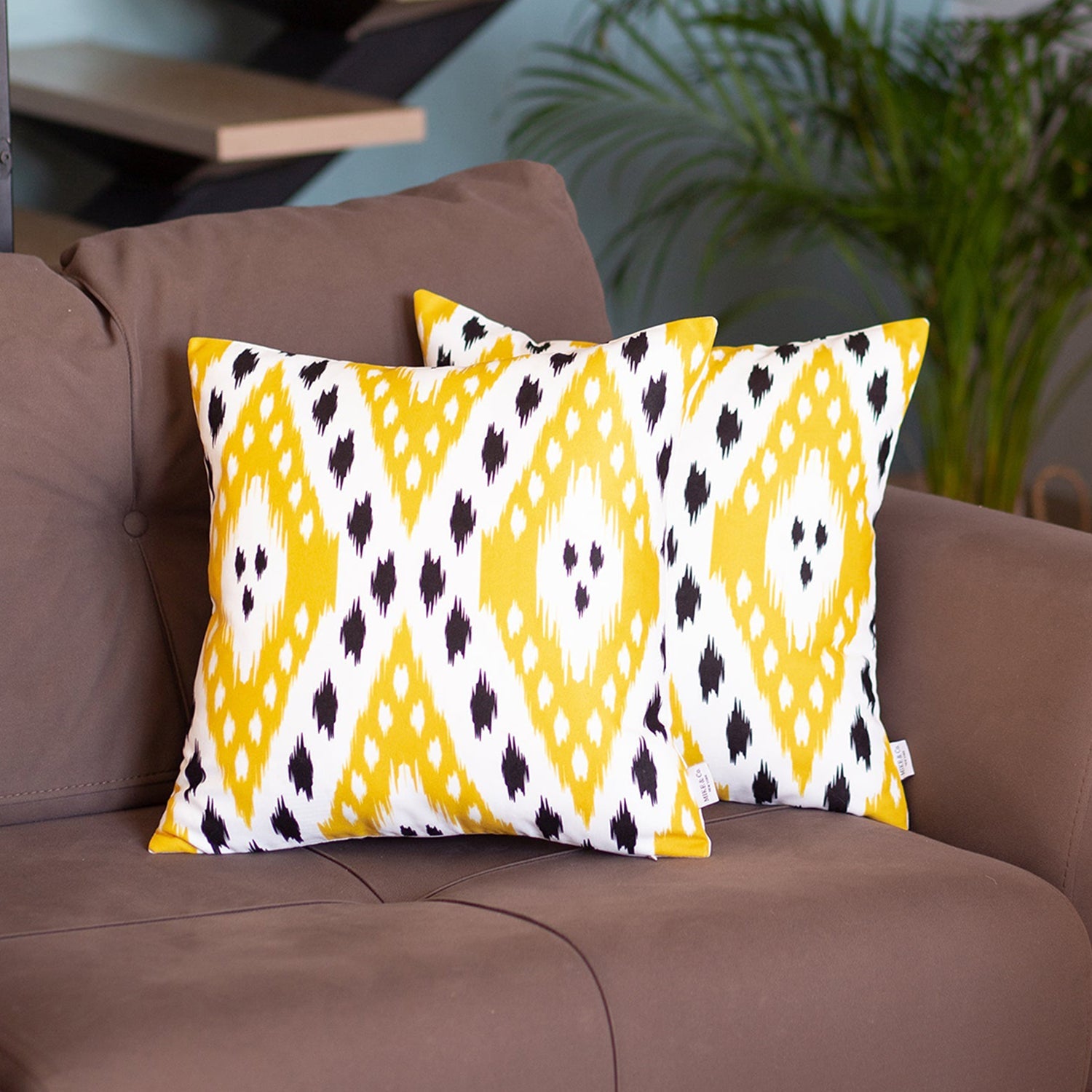 A pair of pillow covers from the Ikat Set of 2 Square 18’’x18’’ Boho Throw Pillow Covers, showcasing a blue and white ikat design with hidden zipper closures, are placed on a brown sofa. In the background, you can see a potted plant and a staircase.