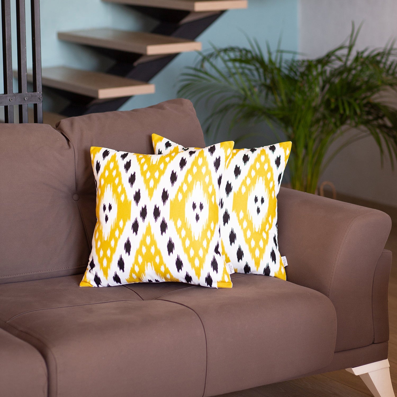 A pair of pillow covers from the Ikat Set of 2 Square 18’’x18’’ Boho Throw Pillow Covers, showcasing a blue and white ikat design with hidden zipper closures, are placed on a brown sofa. In the background, you can see a potted plant and a staircase.