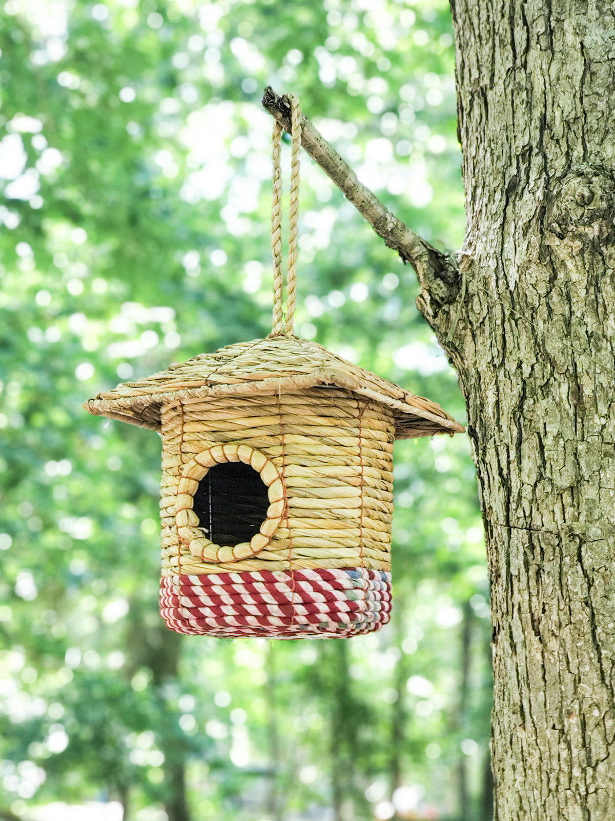 The Seagrass & Sari Birdhouse - Cottage is a handcrafted nest featuring a circular entrance, a red-and-white checkered base, and hangs by a rope loop, making it an environmentally friendly addition to your garden.
