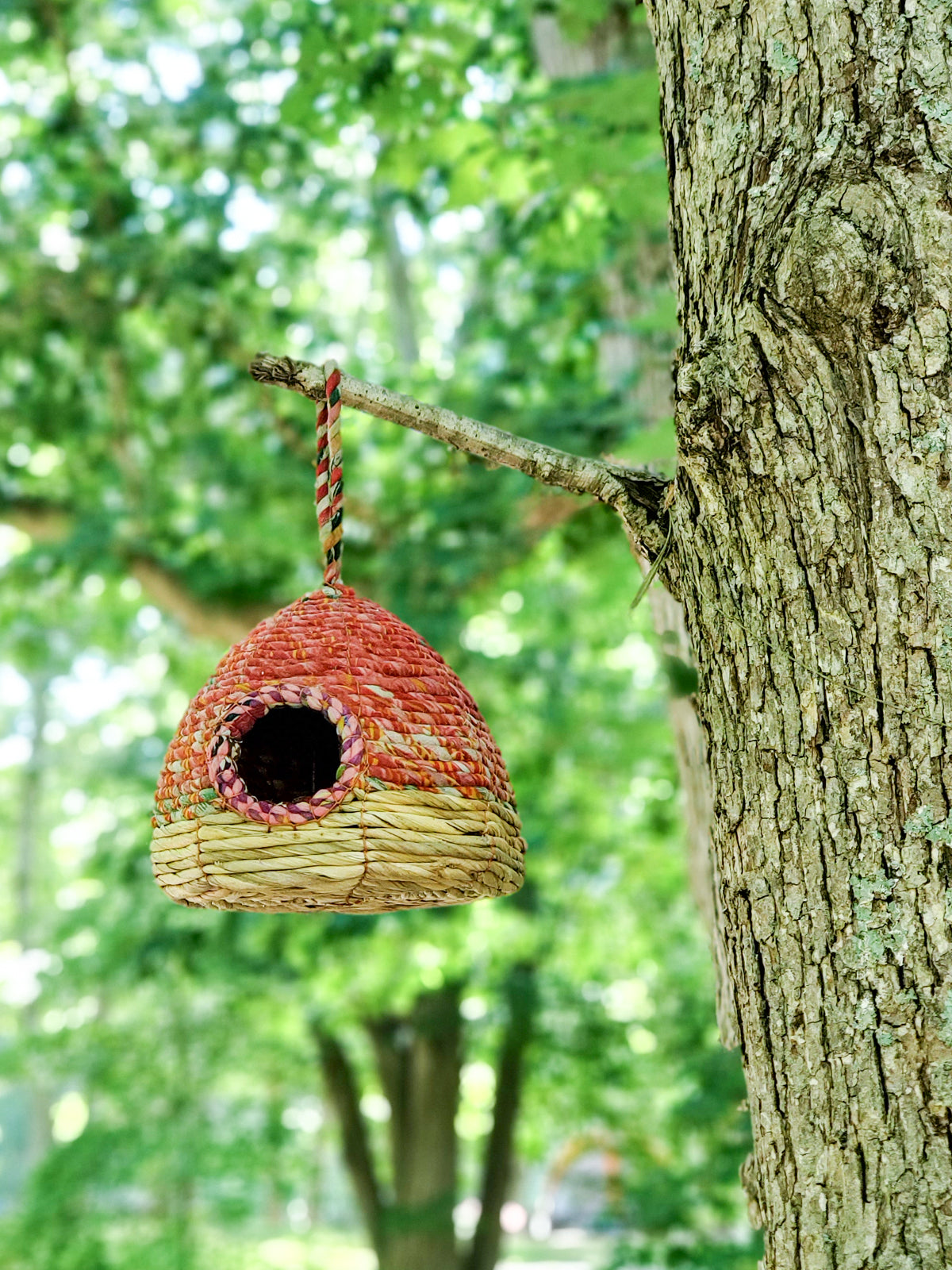 The Seagrass & Sari Birdhouse - Garden is an eco-friendly, handcrafted birdhouse showcasing a woven red and beige design, featuring a circular entrance and a looped handle on top.