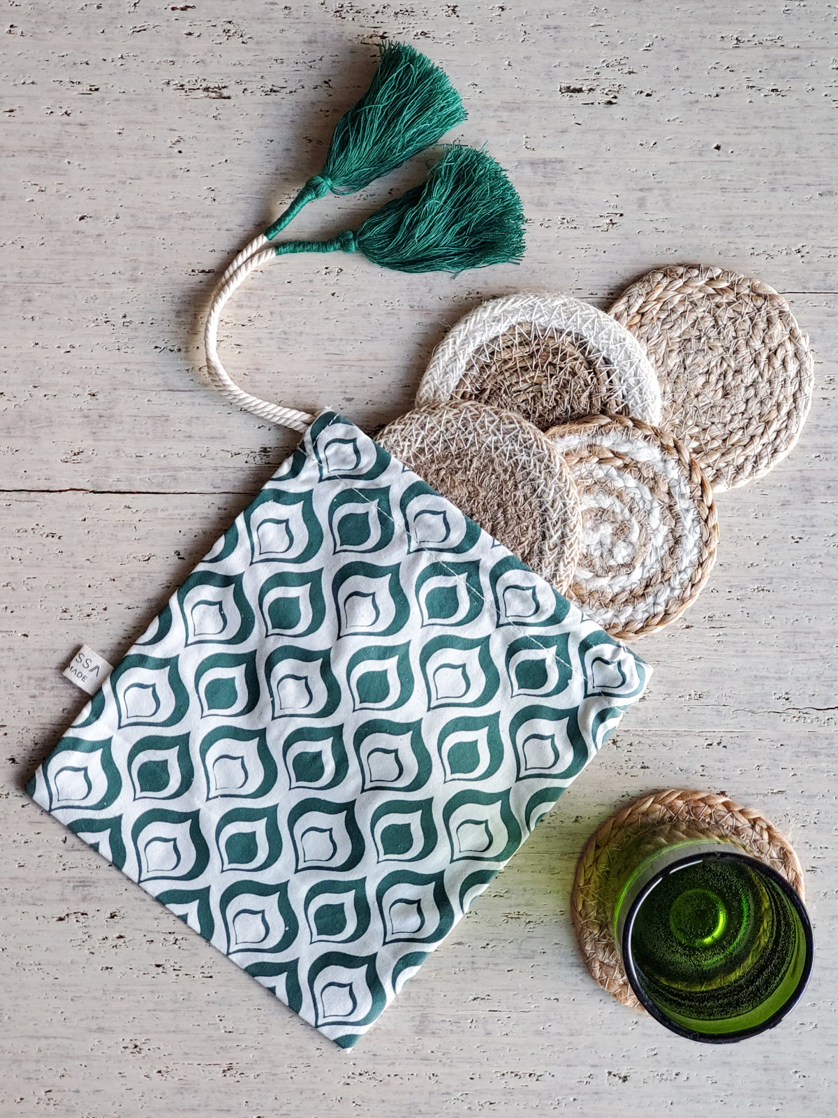 Introducing the Natural Coaster Gift Set with Green Pouch: a sustainable gift featuring a fabric pouch adorned in a green and white pattern, containing six round woven jute coasters. Delightful green tassels add an extra touch of charm, and each piece is skillfully crafted by fair trade artisans.