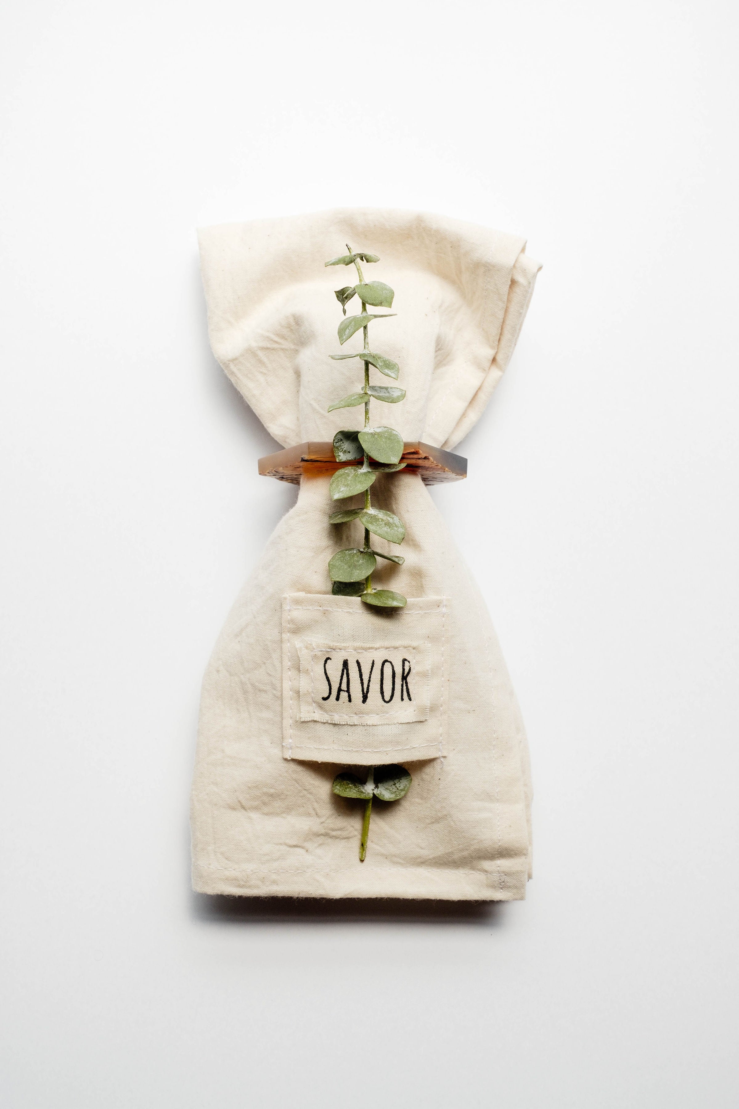 Pocket napkin set of four, skillfully crafted from up-cycled fabric, folded with a eucalyptus sprig and wooden ring, labeled "SAVOR.