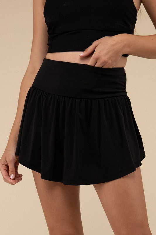 Close-up of a woman in a black crop top and Wide Band Tennis Skirt with Zippered Back Pocket, pulling the skirt to show matching shorts with a zippered back pocket underneath.