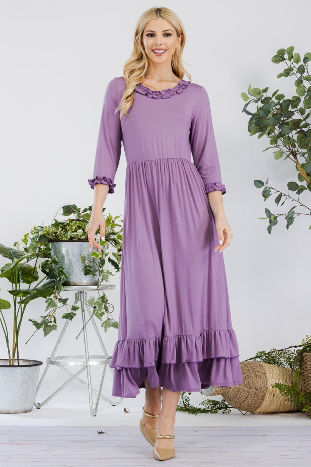 A woman in the Celeste Full Size Layered Ruffle Hem Dress with Pockets, featuring an A-line silhouette and 3/4 sleeves, stands on a wooden floor surrounded by potted plants and a wicker basket.