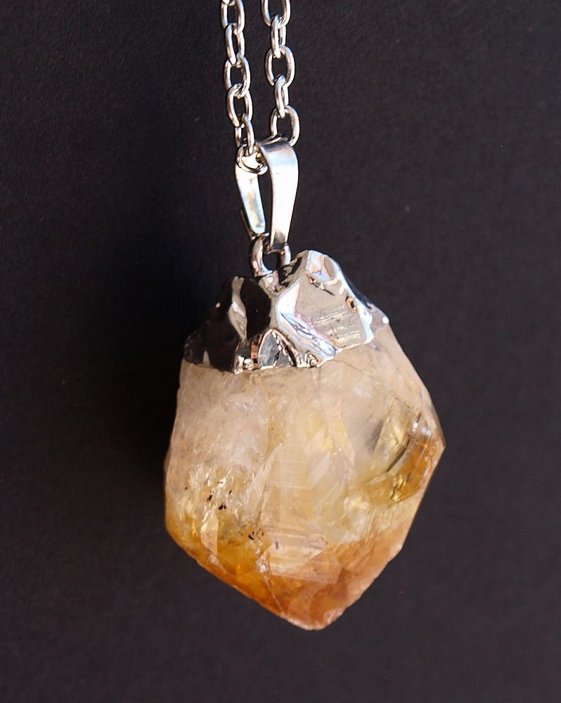 The Raw Citrine Crystal Statement Necklace features a rough citrine crystal and a silver-capped pendant on a shimmering silver chain, elegantly displayed against a black background.