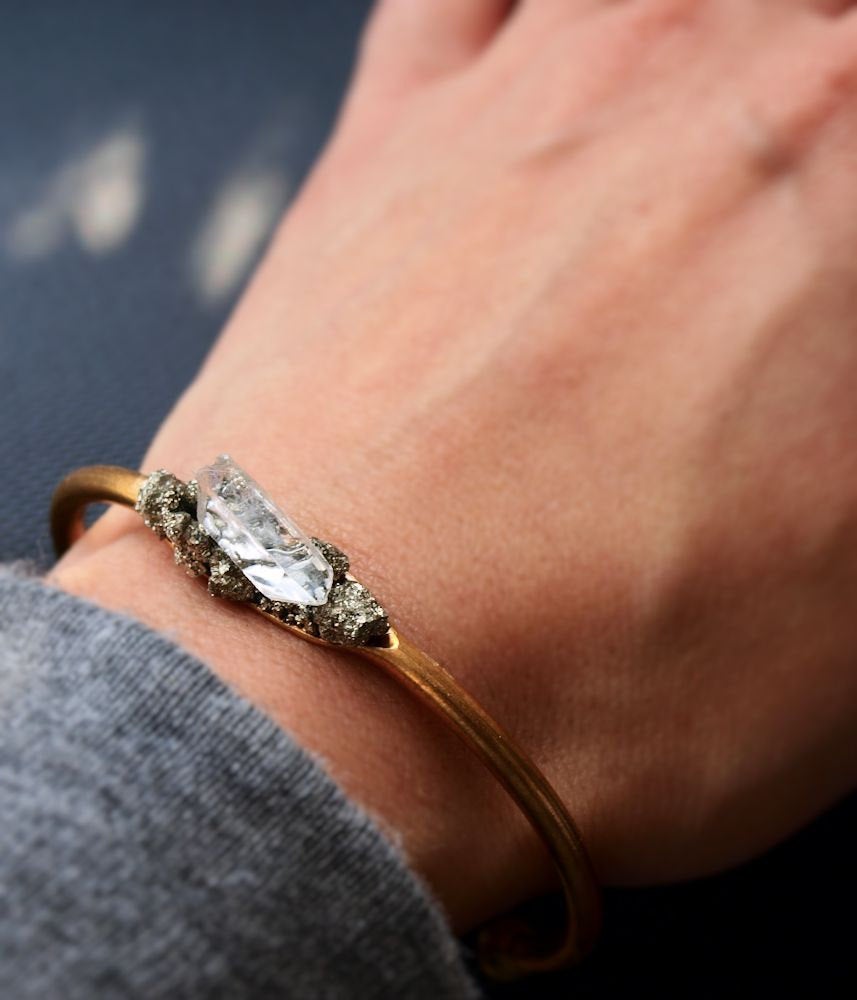 Pyrite and Quartz Gold Bangle, featuring a large rectangular quartz crystal and small round crystals for spiritual protection, set against a black textured background.