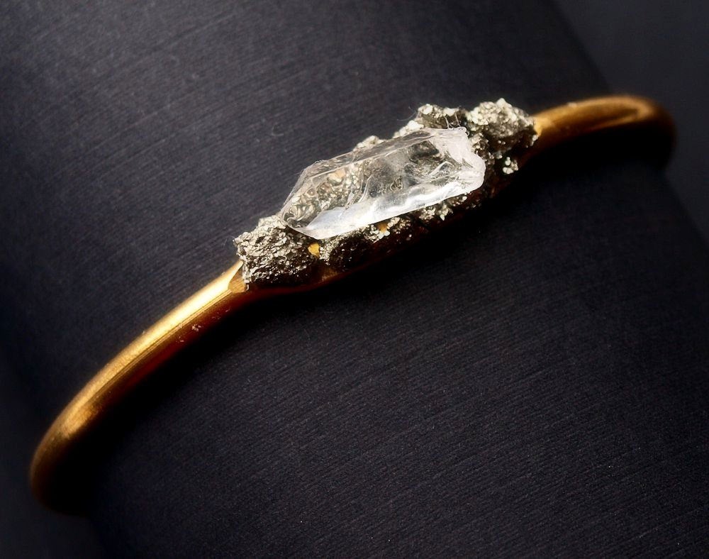 Pyrite and Quartz Gold Bangle, featuring a large rectangular quartz crystal and small round crystals for spiritual protection, set against a black textured background.