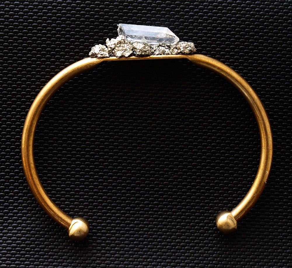 Pyrite and Quartz Gold Bangle, featuring a large rectangular quartz crystal and small round crystals for spiritual protection, set against a black textured background.